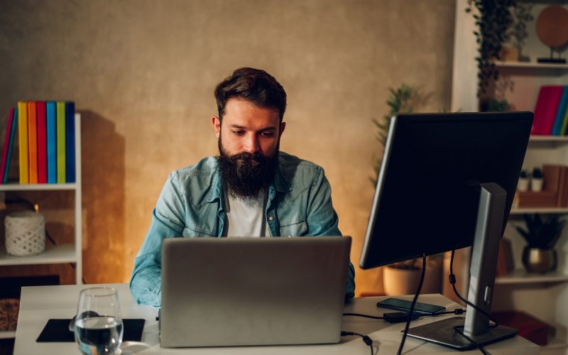 A bearded hipster is typing on a laptop and developing an AI web app on his laptop while sitting in his cozy office at night. A focused senior web app developer is working on an AI generator.